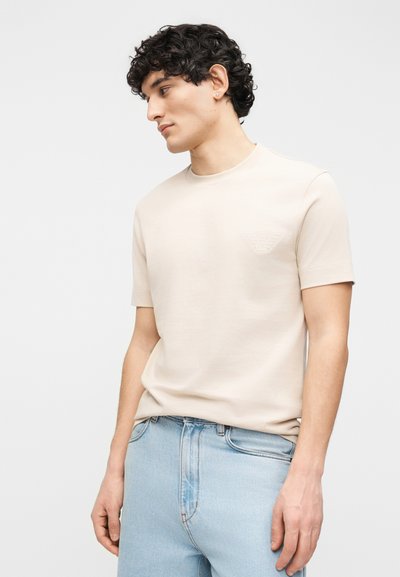 Νεαρός με σγουρά σκουρόχρωμα μαλλιά, φορώντας μπεζ κοντομάνικο πουκάμισο και ανοιχτόχρωμο τζιν, κοιτάζει προς το πλάι σε απλό φόντο.