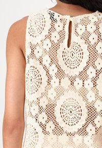 Blonde med blomstermuster laget av kremfarget heklemateriale med sirkulære blomsterdesign og en nøkkelhull-utskjæring bak.