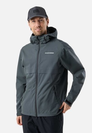 Homme portant une veste à capuche gris foncé et une casquette noire, debout avec une main dans la poche devant un fond blanc.