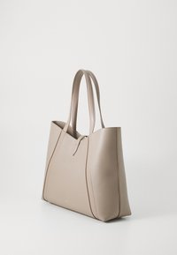Beige leather tote bag with two long handles, angular design, smooth texture, and minimalistic detailing. Features a central closure.