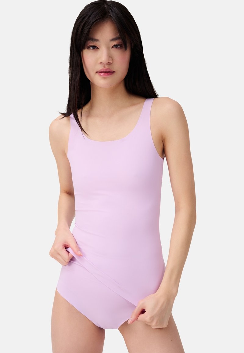 Jonge vrouw met lang, donker haar die een lichtpaarse mouwloze bodysuit draagt en de onderrand met haar handen aanpast.