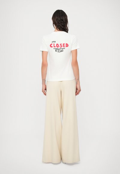 Biele tričko s červeným grafickým nápisom "ZATVORENÉ" na chrbte, kombinované s krémovými širokými nohavicami, predstavujúce neformálny outfit.