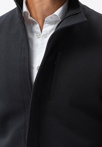 Svart zip-up jacka med texturerad tyg och ståkrage, med en dold zip och en bröstficka. Buren över en vit skjorta.