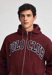 Vinröd zip-up hoodie i mjukt tyg, med en stor "POLO CLUB"-logotyp i vitt och marinblått över bröstet, och justerbar huva.