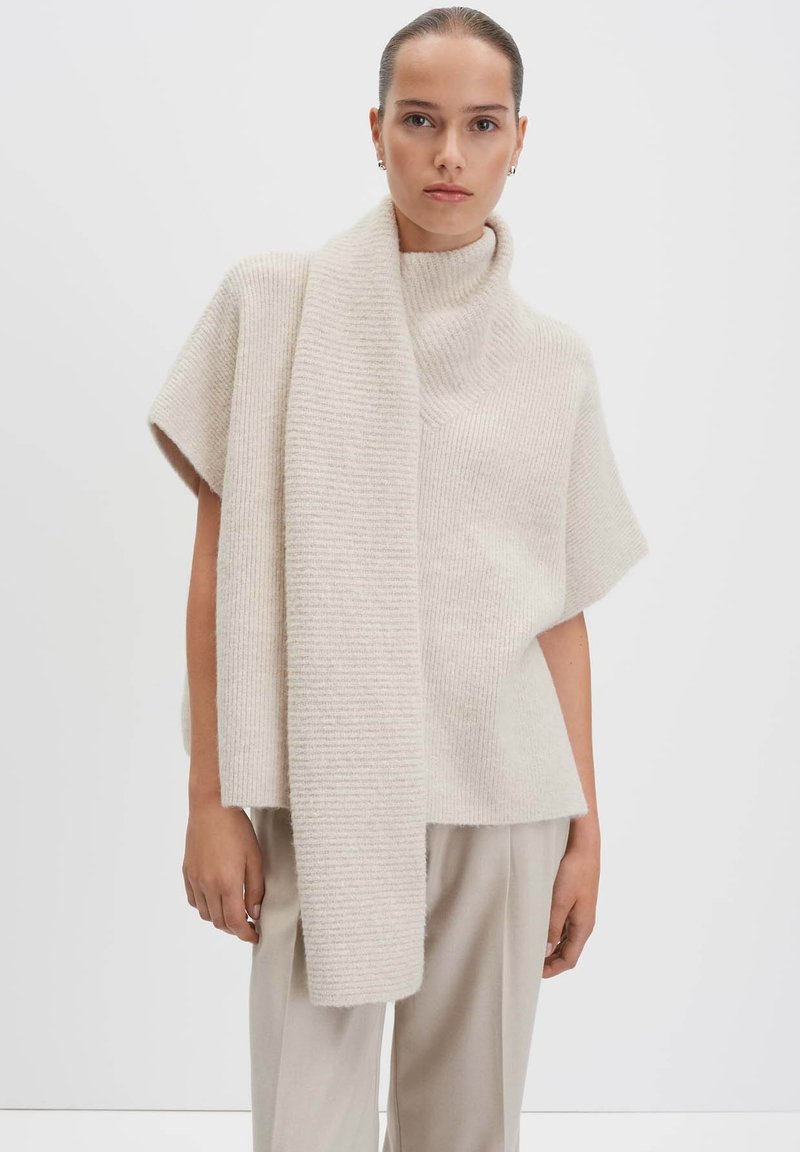 Donna che indossa un maglione lavorato a maglia color crema con maniche corte e una sciarpa lunga attaccata, abbinato a pantaloni beige chiaro, su uno sfondo neutro.