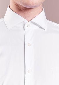 Camisa blanca abotonada hecha de tela suave, con cuello puntiagudo y pequeño detalle bordado en el pecho. Cinco botones visibles.