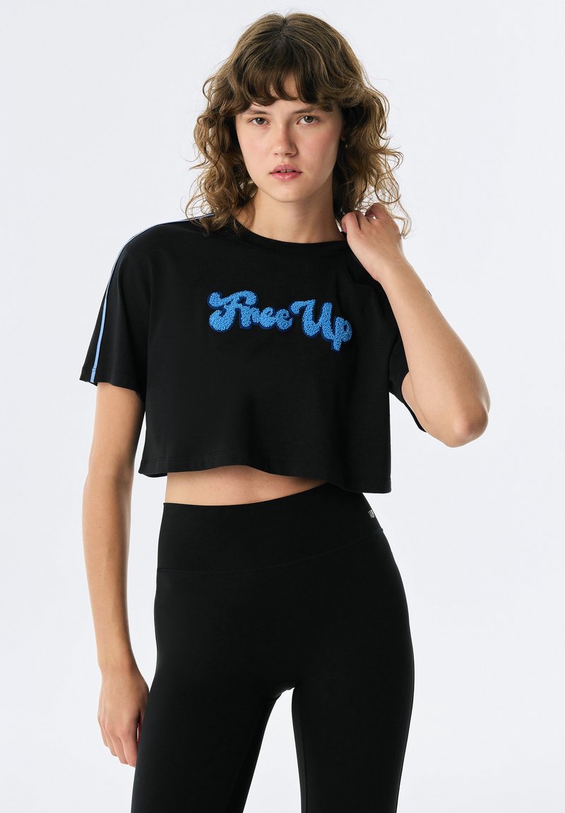 Zwarte cropped t-shirt met een blauwe sprankelende "Rise Up" graphic, korte mouwen met lichtblauwe accenten, gecombineerd met zwarte leggings.