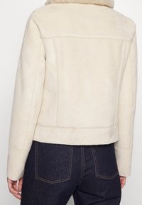 Giacca in suede beige con colletto peloso, design corto e dettagli cuciti, abbinata a jeans scuri con tasche posteriori.