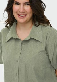 Žalia kordūroja mygtukinė marškena su smailiu apykakle, trumpomis rankovėmis ir subtilia briaunota tekstūra. Viena kišenė kairėje pusėje.