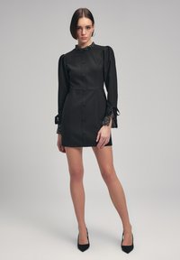 Robe noire avec de délicates manches bouffantes, fermeture à boutons et col perlé. Présente des poignets transparents à motifs et une silhouette ajustée.