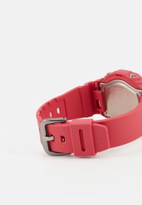Rdeča silikonska ura z zaponko iz kovine, ki se ponaša z gladko teksturo in nastavljivo dolžino. Oblika vključuje izreze za zračnost.