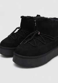 Botines de ante negro con una suela plataforma gruesa negra, forrados con piel sintética, con cordones delanteros y cierre de cremallera en el lateral.