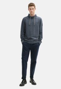Sweatshirt van donkergrijze gebreide stof met een hoge hals, trekkoord en subtiele logo; gedragen met marineblauwe broek en zwarte veterschoenen.