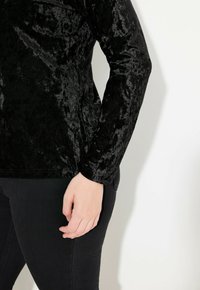 Schwarzes Samtoberteil mit langen Ärmeln und glänzender Textur, figurbetontem Design, abgerundetem Saum und glatter Oberfläche, kombiniert mit schwarzen, eng anliegenden Hosen.