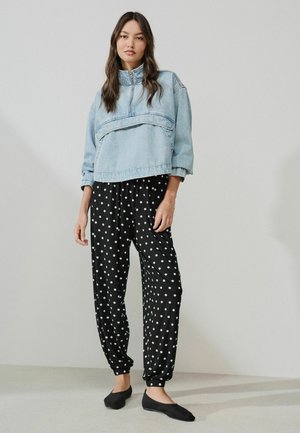 Vrouw gekleed in een lichtblauwe cropped denim jas en zwarte losse broek met witte polka dots, staand tegen een effen muur.