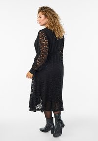 Čierne čipkované šaty s dlhými rukávmi, priliehavým pásom a rozšíreným lemom. Kombinované s čiernymi členkovými čižmami, ktoré majú textúrovaný vzor.