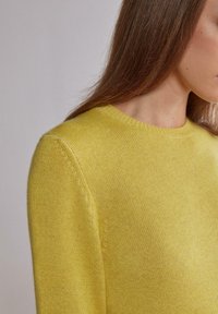 Femme aux cheveux bruns et raides portant un pull en laine jaune clair, montrée de l'épaule jusqu'au bas du visage sur un fond neutre.