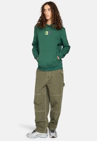 Sudadera de algodón verde con un bolsillo frontal y detalle gráfico. Combinada con pantalones khaki holgados que presentan acentos de costura. Lleva zapatillas gris.