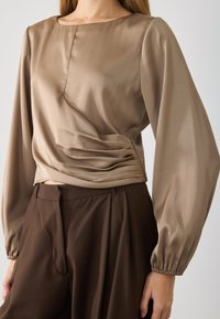 Beige satijnen blouse met lange pofmouwen, voorzien van een zijruche-detail en een ronde halslijn, gecombineerd met bruine hoog-gerokte broek.