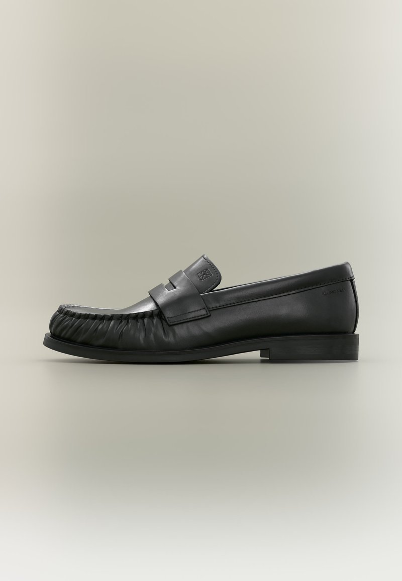 Mocassin penny en cuir noir avec détail plissé au bout et talon bas empilé, vu de côté sur un fond neutre.