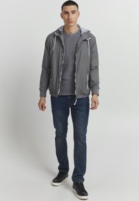 Lett grå zip-up jakke med hætte, der har hvide snore, kombineret med en struktureret grå sweater, blå jeans og sorte sneakers.