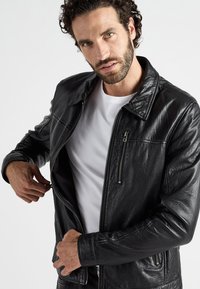 Zwarte leren jas met een rits aan de voorkant, staande kraag en twee ritszakken. De textuur lijkt iets gekreukt en soepel.