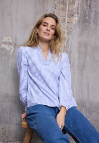 Blouse rayée bleu et blanc avec un col en V et un bord à volants, manches longues avec poignets froncés, portée avec un jean bleu. Fond chaise en bois.