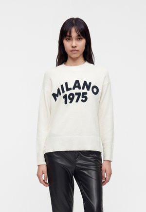Femme aux cheveux bruns raides porte un pull blanc imprimé de "MILANO 1975" et un pantalon en cuir noir, debout devant un fond uni.