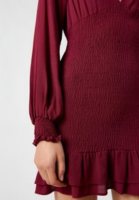 Vestido burdeos con largas mangas abullonadas. Presenta un acabado arrugado y un dobladillo con volantes. Está hecho de una tela ligera con un acabado suave.