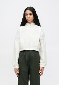 Κοντό μπεζ σακάκι με ψηλό γιακά, κλείσιμο με φερμουάρ και ελαστικές μανσέτες, συνδυασμένο με σκούρο πράσινο παντελόνι με πλαϊνές τσέπες.