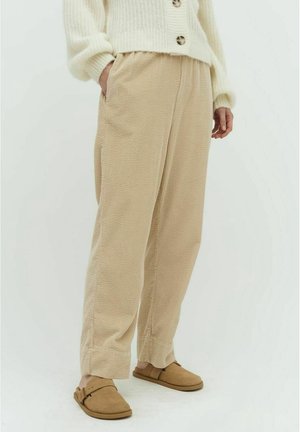 Osoba w beżowych spodniach o fakturze, beżowych butach wsuwanych i kremowym swetrze z guzikami, stojąca z jedną ręką w kieszeni.