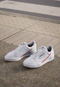 White leather sneakers with red and blue accents, perforated toe box, flat rubber sole, and Adidas branding on the side.