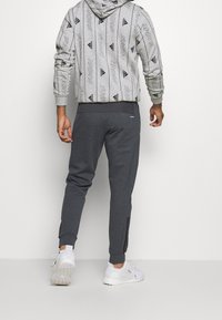 Grauer Hoodie mit schwarzen Logos, kombiniert mit dunkelgrauen Jogginghosen in Slim Fit und seitlichen Reißverschlüssen, sowie weißen Sportschuhen.