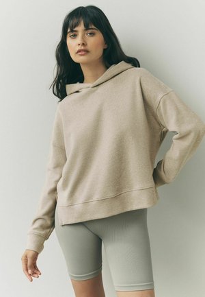 Beżowy krótki hoodie z kapturem na sznurku i prążkowanymi rękawami, w połączeniu z jasnoszarymi prążkowanymi szortami rowerowymi. Miękki, luźny krój.