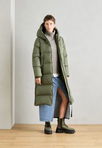 Abrigo de plumas verde oliva con capucha, de tela texturizada, de longitud larga, llevado sobre un suéter gris y una falda vaquera con una abertura lateral, combinado con botas de combate.