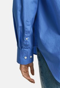 Langarmige blauwe shirt van gladde stof, met witte knopen op de manchetten en subtiele tonale stikdetails langs de mouw.