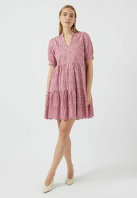 Pink cotton dress with a v-neckline, short puff sleeves, eyelet embroidery, tiered design, and scalloped hem. Worn with beige heels.