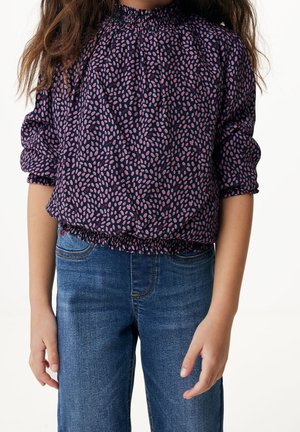 Barn iført en marineblå bluse med lyserøde bladmønstre og blå denimjeans, stående mod en hvid baggrund.