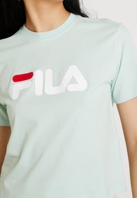 Camiseta de algodón azul claro con un destacado logo de FILA en blanco y rojo en el pecho. Mangas cortas y escote redondo.