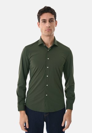 Uomo con capelli corti e scuri che indossa una camicia verde scuro aderente a maniche lunghe con bottoni e pantaloni blu scuro, in piedi contro uno sfondo bianco.