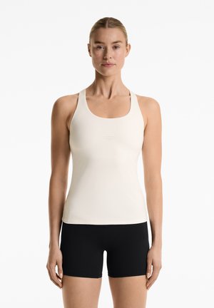 Lys beige tanktop med rund halsudskæring og racerback-design, der har perforeret detaljer i siderne, parret med sorte shorts.