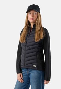Jeune femme portant une veste matelassée noire, un sous-pull noir à manches longues, un jean bleu et une casquette de baseball noire, debout devant un fond blanc.