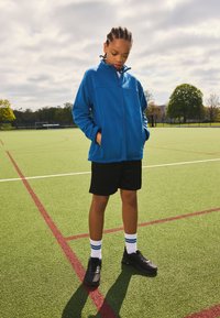 Blå fleecejakke med lynlås, sorte shorts, sorte sportssko og hvide sokker med blå striber, stående på en grøn kunstgræsbane.