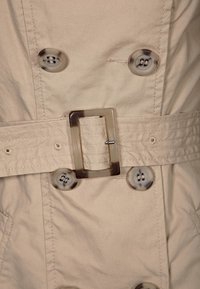 Beige trench coat med midja som är bältad, stora knappar och en rektangulär genomskinlig spänne. Tyget har en slät, strukturerad yta.