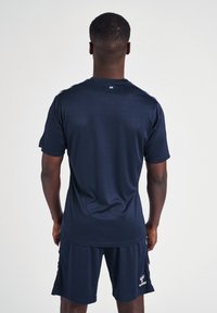 Homme portant une tenue de sport bleu marine à manches courtes et short, debout de dos sur un fond uni.