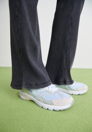 Persona vistiendo pantalones negros acanalados y zapatillas azul claro, de pie sobre un suelo alfombrado verde.