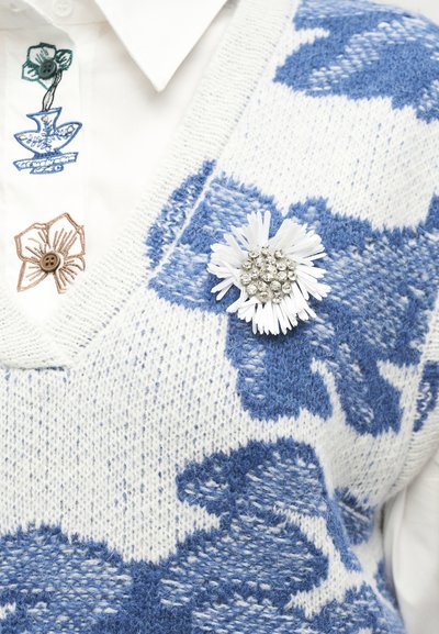 Chemise blanche avec des fleurs brodées et un gilet tricoté blanc et bleu avec une broche florale blanche avec un centre en cristal.