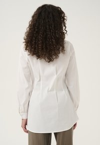 Camicia bianca a maniche lunghe con vestibilità rilassata, polsini arricciati e un orlo posteriore più lungo. Realizzata in tessuto liscio con una leggera trama.