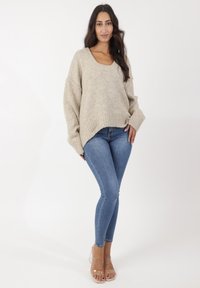 Suéter de punto beige de gran tamaño con escote en forma de barco y dobladillo acanalado, combinado con jeans skinny azul y tacones transparentes.