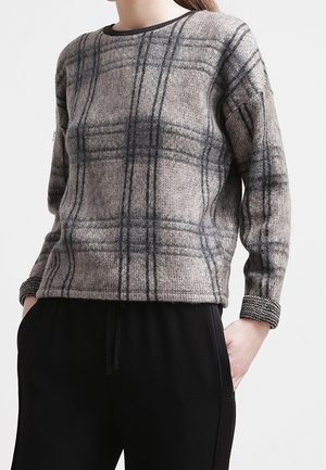 Femme portant un pull à manches longues à carreaux gris avec des poignets noirs et un pantalon noir, debout les mains dans les poches devant un fond uni.